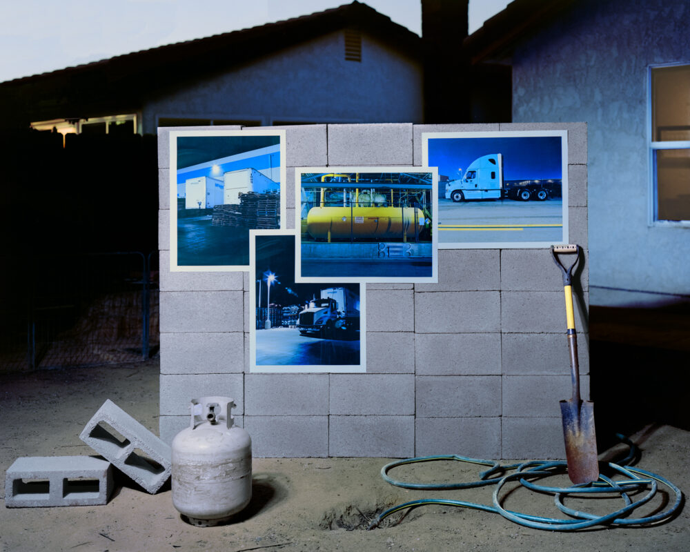 Great block wall in a suburban backyard setting with four photosgraphs attached to it, two of semi-trucks, one of a warehouse, and one of a large propane fuel tank. leaning on the wall to the right is a small shovel. On the ground to the left is a small propane tank. A garden hose lays on the ground in front of the wall.
