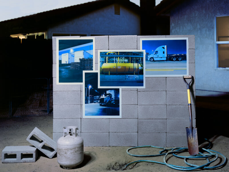 Great block wall in a suburban backyard setting with four photosgraphs attached to it, two of semi-trucks, one of a warehouse, and one of a large propane fuel tank. leaning on the wall to the right is a small shovel. On the ground to the left is a small propane tank. A garden hose lays on the ground in front of the wall.