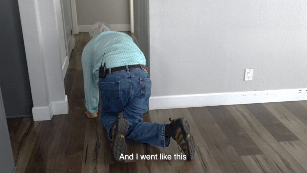 older man in blue jeans and and lite blue striped long sleeved shirt crawling on the dark wood floor through an interior doorway of a home