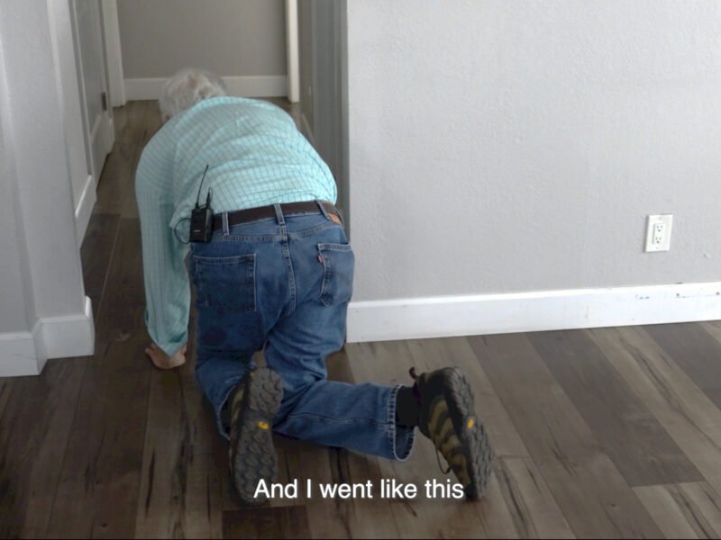 older man in blue jeans and and lite blue striped long sleeved shirt crawling on the dark wood floor through an interior doorway of a home