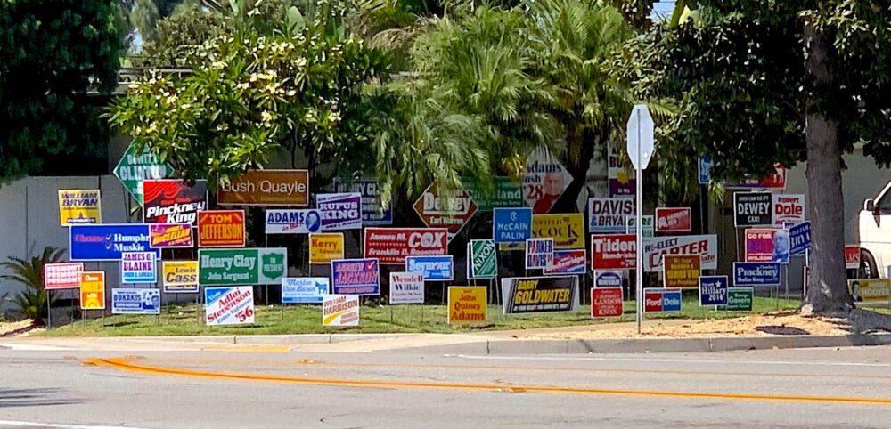 Front yard of home located on street corner with 58 colorful yard signs installed on the front lawn. Each sign bearing the name of former candidates for US President that lost their election.