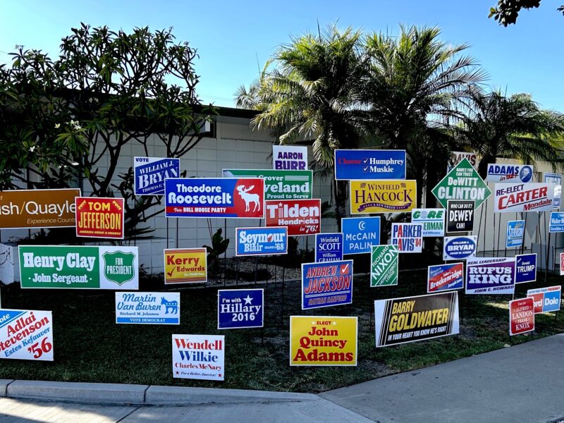 Front yard of home located on street corner with 58 colorful yard signs installed on the front lawn. Each sign bearing the name of former candidates for US President that lost their election.